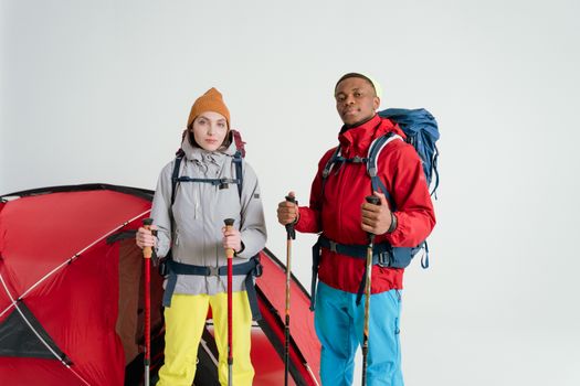 Two adults equipped with camping gear and hiking sticks pose in front of a tent on a white background.