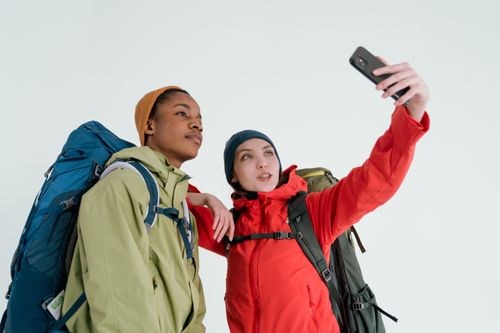 Free A diverse couple in jackets and backpacks taking a selfie in a studio setting, capturing their travel spirit. Stock Photo
