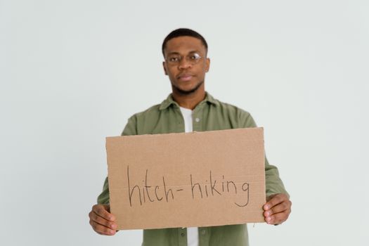 Portrait of a man holding a cardboard sign for hitchhiking, standing against a neutral background.