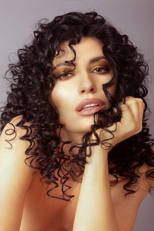 Free Alluring female with makeup and curly black hair looking at camera while sitting on gray background with bare shoulders in studio Stock Photo