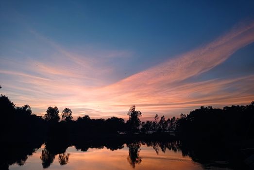 Beautiful sunset reflecting over a serene lake with silhouetted trees in Dhaka, Bangladesh.