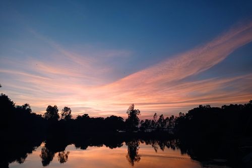 Free Beautiful sunset reflecting over a serene lake with silhouetted trees in Dhaka, Bangladesh. Stock Photo