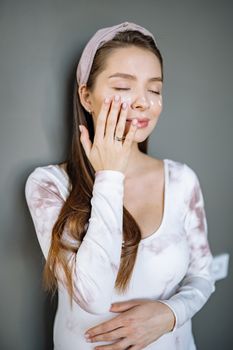 A young woman in casual attire enjoys a skincare routine applying cream to her face.