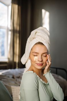 A serene woman with a towel on her head enjoys skincare with under eye masks indoors.