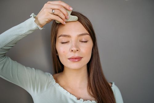 Free A woman relaxes with a jade gua sha tool for a facial skincare routine against a gray background. Stock Photo