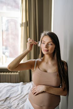 A serene pregnant woman in a morning skincare routine with a jade roller by a bright window.