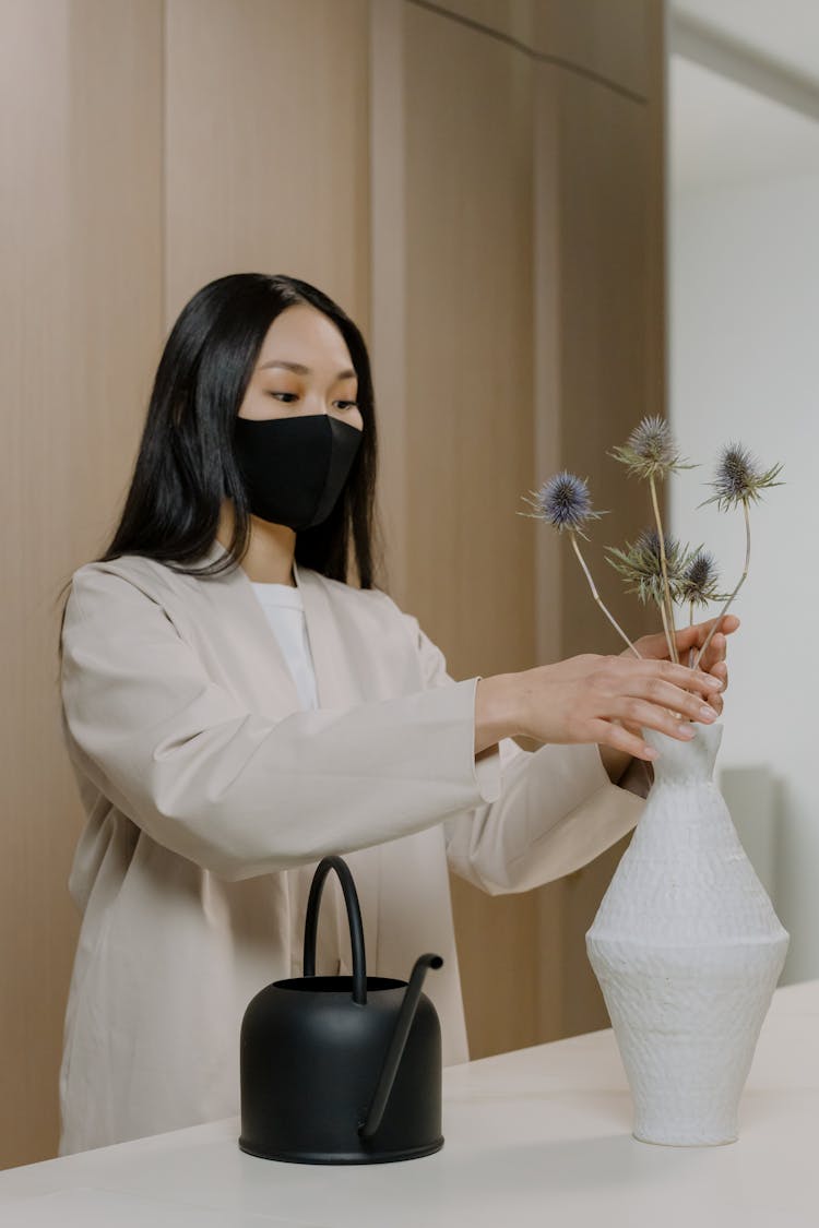 Woman With Face Mask Arranging Flowers In White Ceramic Vase