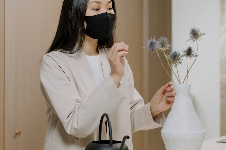 Woman In Beige Coat Wearing Black Face Mask Arranging Flowers In A Vase