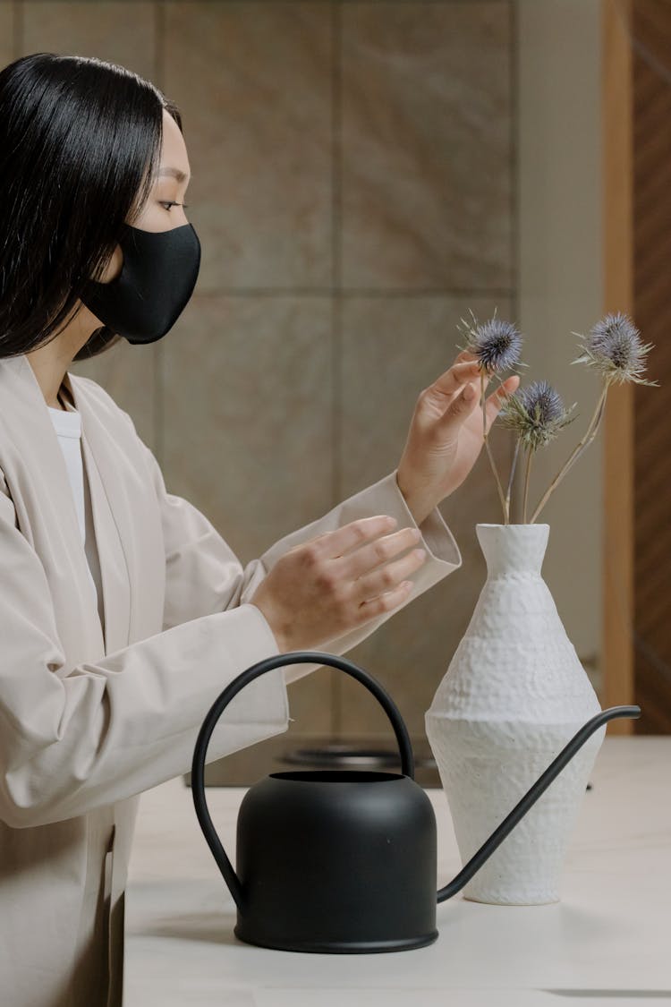 Woman In Beige Coat Wearing Black Face Mask Arranging Flowers In A Vase