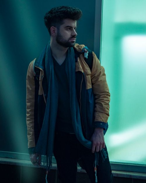 Free Portrait of a fashionable man with a full beard, wearing a scarf and jacket, standing in a modern urban setting. Stock Photo