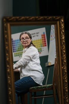 Senior woman reflected in mirror, seated with a periodic table in view, suggesting a science theme.
