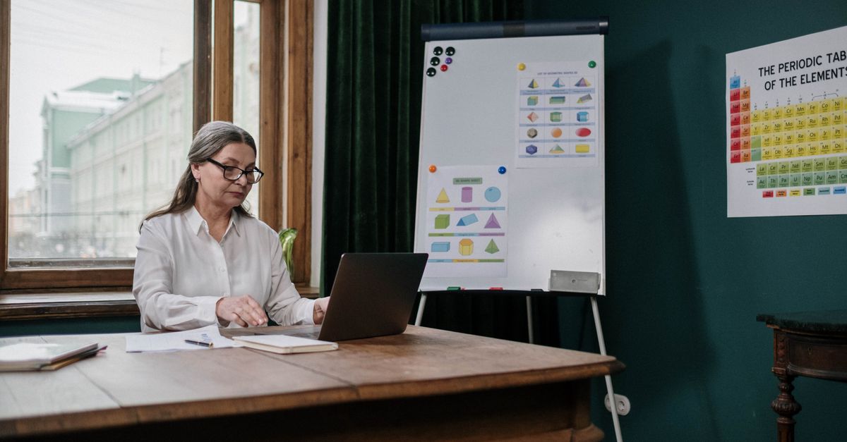 Elderly woman in classroom using laptop with periodic table and charts.