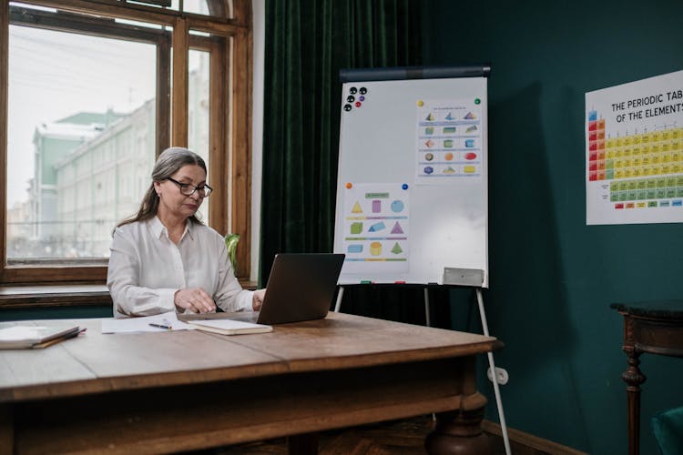 Elderly Woman Sitting At A Wooden Desk Using A Laptop 