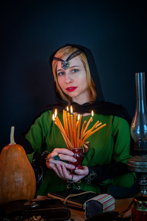 Young female soothsayer with talisman on head and flaming candles looking at camera against decorative pumpkin