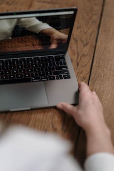 Close-up of a person using a laptop on a wooden table, focusing on reflection and hand.