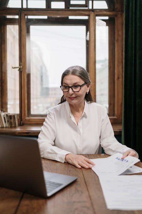 A Woman in White Long Sleeves Looking at a Laptop while Holding Papers