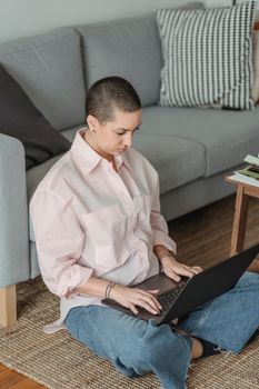 From above of young concentrated female remote worker with short hair in casual clothes typing on laptop while sitting on floor near sofa at home