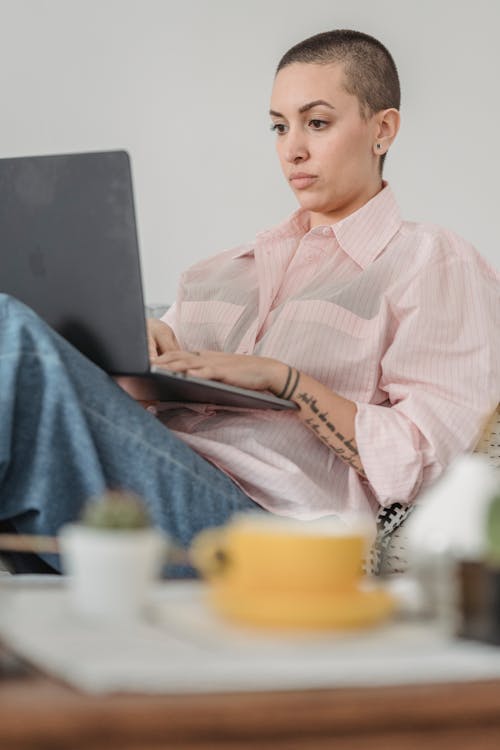 Serious young tattooed female freelancer with shaved head in casual clothes sitting on sofa and typing on netbook while working remotely from home