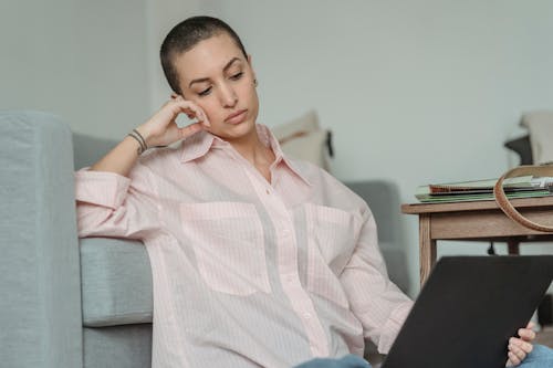 Pensive young self employed woman with short dark hair in casual clothes leaning on hand while sitting on floor near sofa during remote work on laptop at home