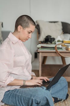 Side view of busy young female freelancer with short hair in casual outfit sitting on floor with crossed legs and browsing laptop while working remotely in light living room