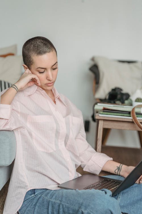 Calm young self employed woman with short dark hair in casual outfit leaning on hand while working distantly on laptop sitting near sofa living room