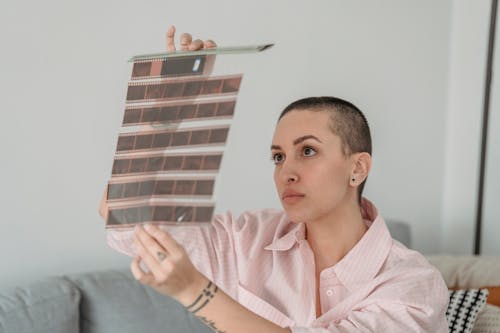 Focused young female photographer with shaved head in stylish shirt examining film tapes while sitting on comfy sofa in modern living room