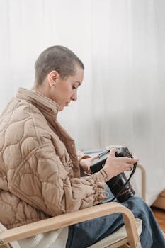 A bald woman in a quilted jacket examines her camera settings while sitting indoors.