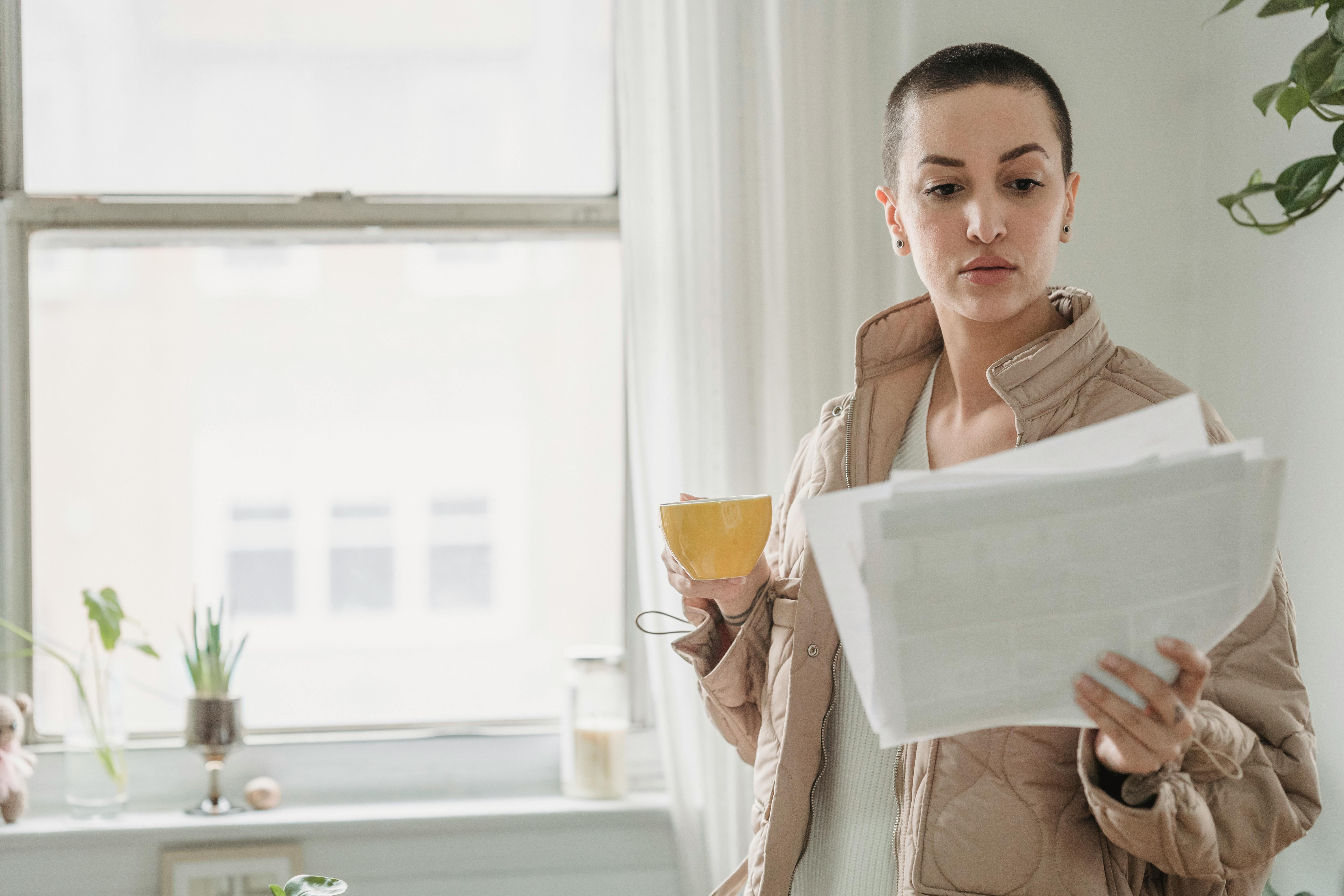 A woman stands by a window holding papers and a coffee cup, in daylight.