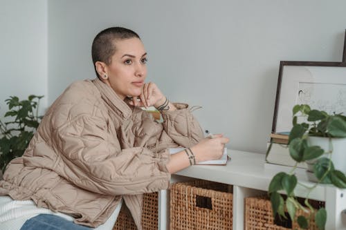 Free Contemplative female writer with tattoos and short hair leaning on desk with agenda while looking away in house Stock Photo