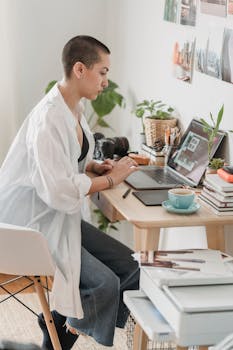 Young woman concentrating on digital work at a home office desk with laptop and camera, surrounded by plants.