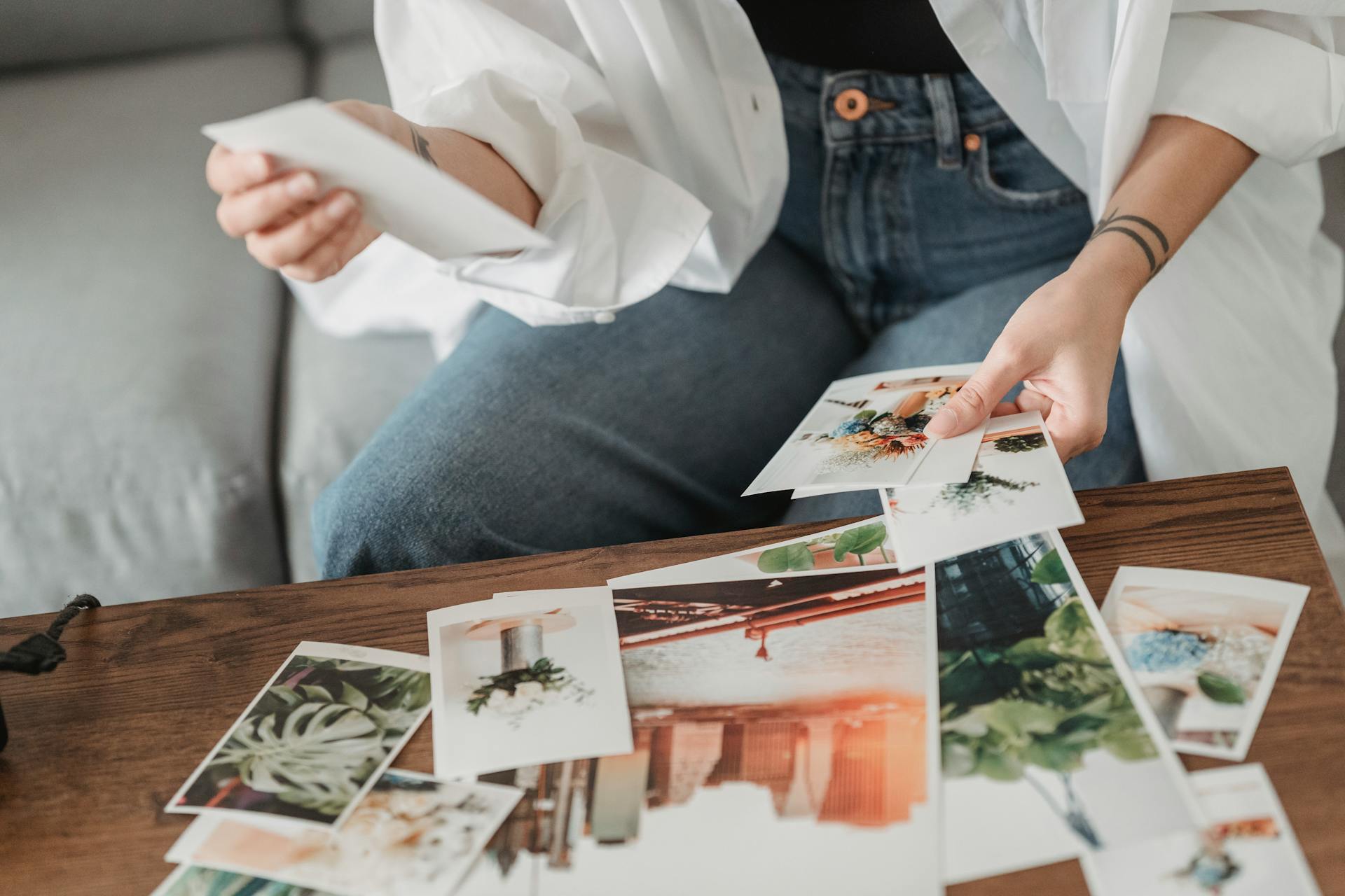 Crop unrecognizable tattooed female sitting on sofa at desk with assorted photos of flowers and town in house room