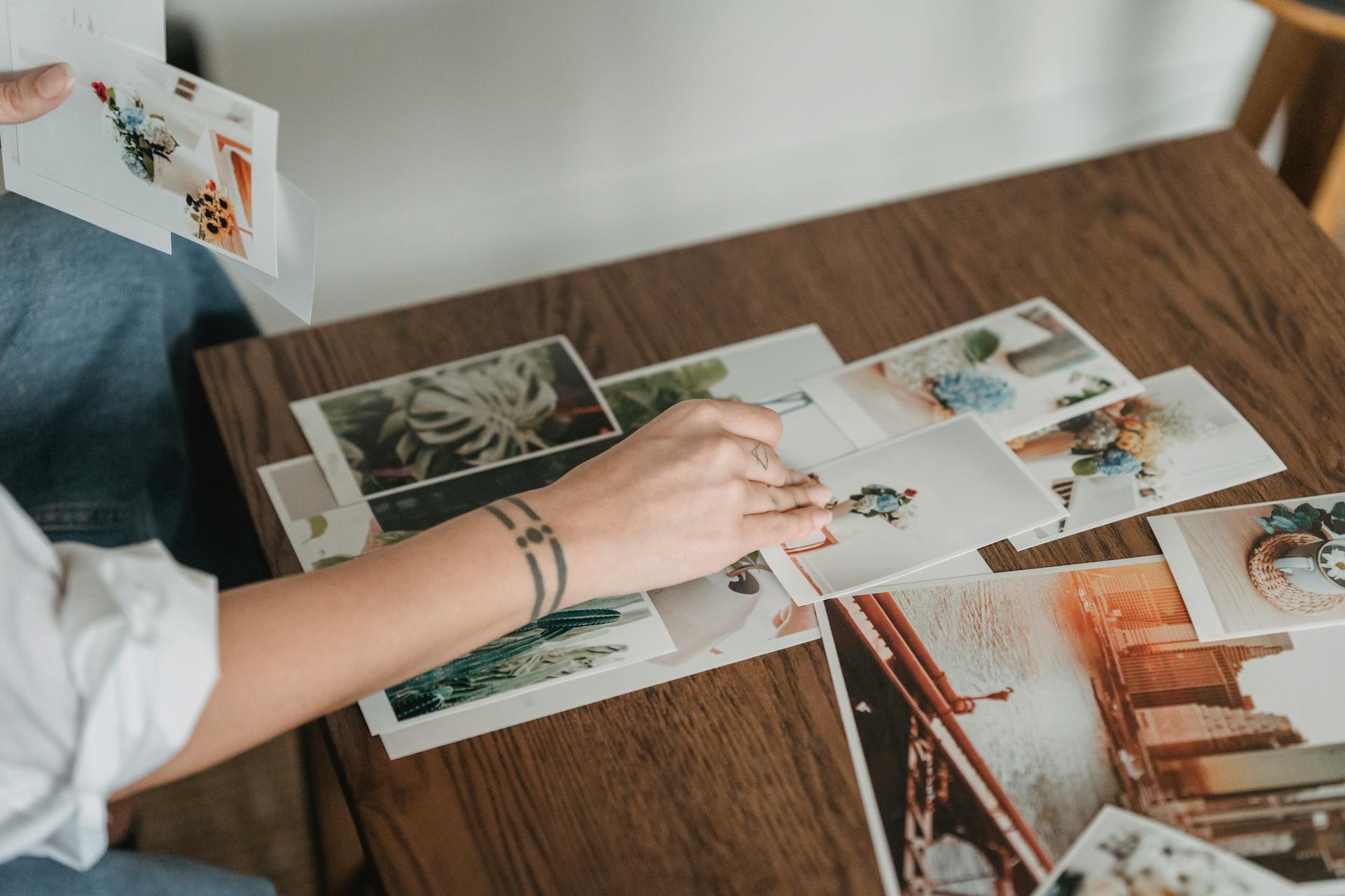 Crop anonymous tattooed female with different photos of flowers and city at desk in house room