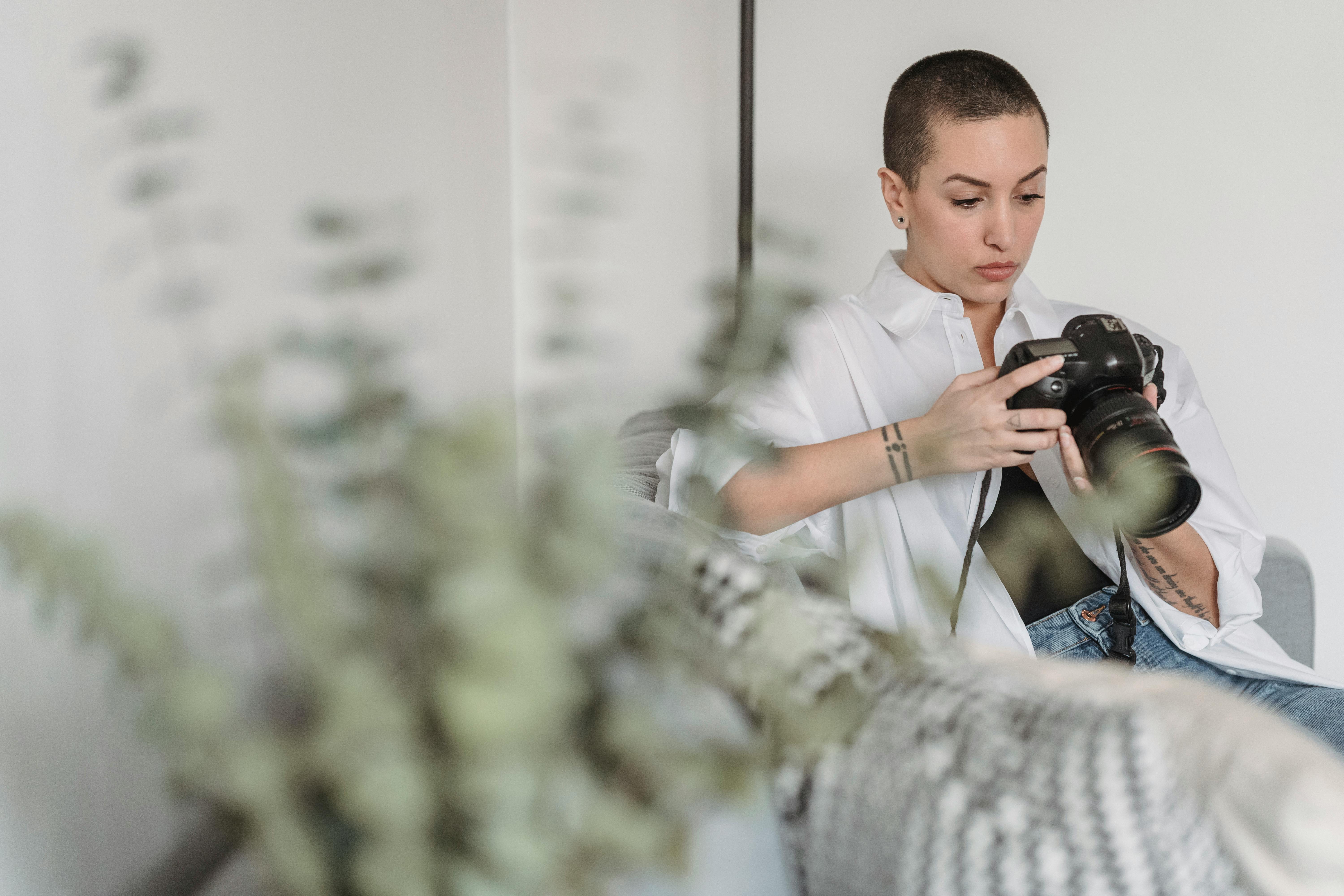 Young tattooed woman with short hair watching digital photo camera while sitting on sofa in house room