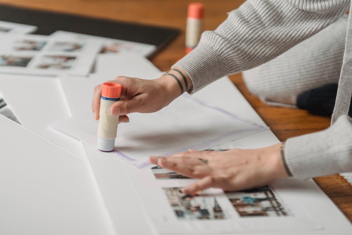 Free Crop anonymous person in warm gray wear applying glue stick on papers and arranging photo album on floor Stock Photo