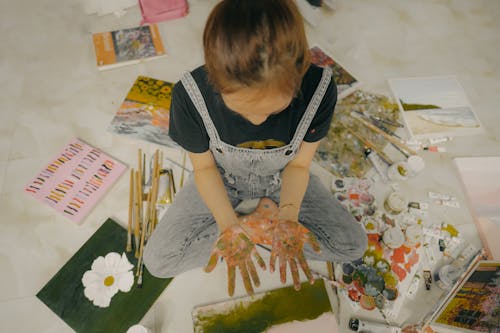 From above crop anonymous girl in casual jumpsuit demonstrating dirty hands while sitting on floor near paints paintbrushes and drawings