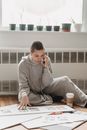 Concentrated young female in gray domestic suit making photo album and having phone conversation while sitting on floor in light room