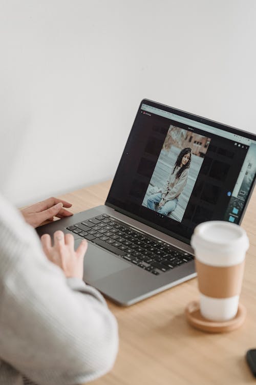 Crop unrecognizable person looking at image on netbook screen while sitting at desk with takeaway coffee