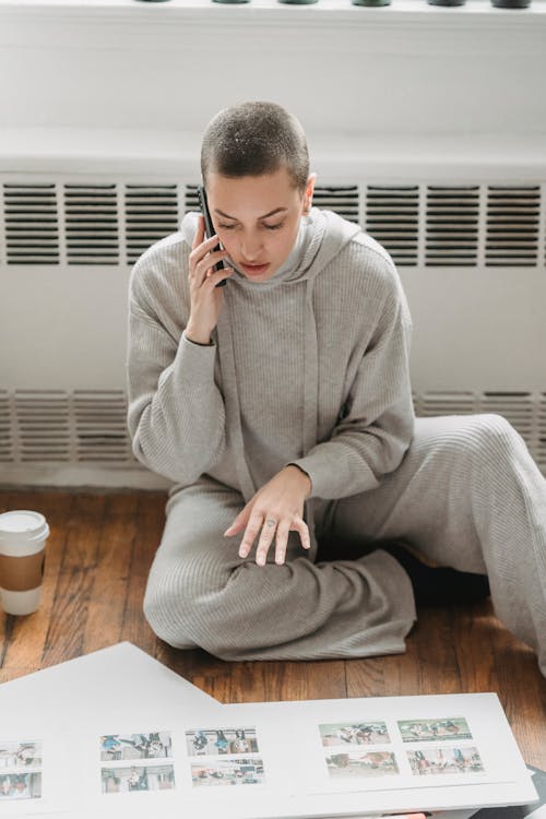 Concerned female in pajama sitting on floor near window while looking down at photos in album