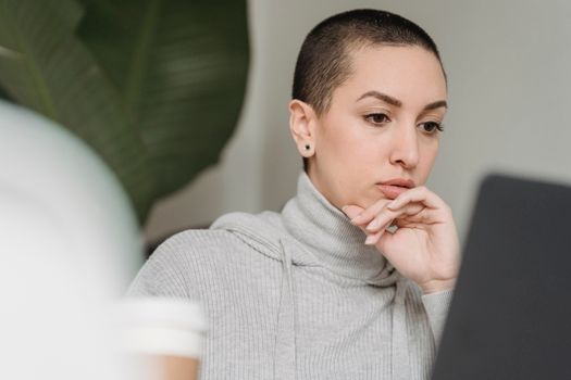 Thoughtful female with short hairstyle looking at netbook screen and touching chin in light room