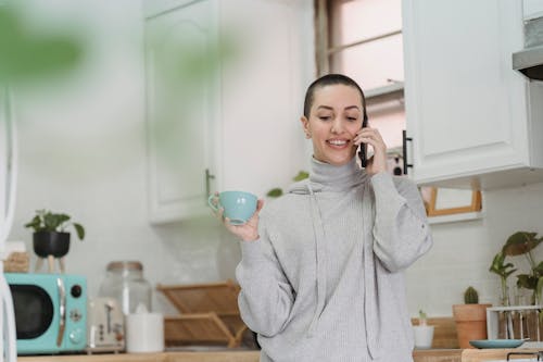 Smiling young female in gray comfy clothes having pleasant conversation on mobile phone and enjoying hot drink while standing in modern kitchen