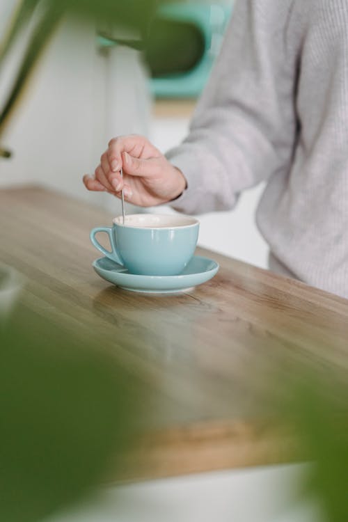 Free Crop anonymous female in gray sweater stirring sugar in cup of freshly brewed coffee while standing near kitchen counter Stock Photo