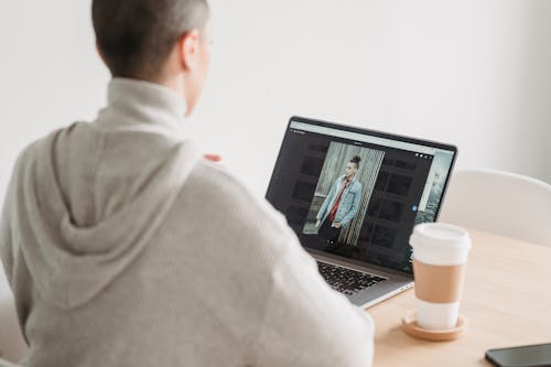 Back view anonymous female in gray hoodie looking at image on netbook screen and sitting at desk with takeaway coffee