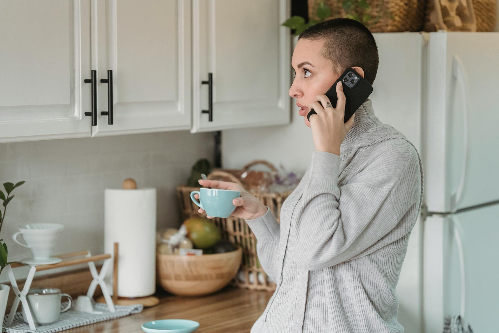 Side view unemotional female with short hairstyle having conversation via mobile phone and enjoying fresh hot drink while standing in modern kitchen