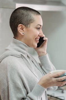 Young woman with short hair smiling during a phone call while holding a cup indoors.