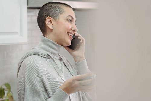 Free Side view cheerful young female in gray turtleneck having phone conversation and enjoying cup of coffee while standing in light kitchen Stock Photo
