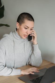 Concentrated young female in gray domestic clothes browsing netbook and having conversation via cellphone in light room