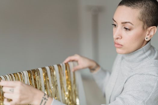 Crop emotionless female in gray wear hanging shimmering golden tinsel on white canvas in light room