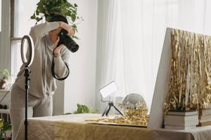 Female photographer with short hair standing at table with shimmering tinsel and party ball near luminous lamp on tripod