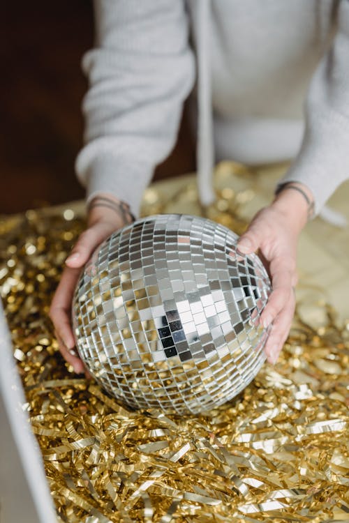High angle of crop anonymous female in casual hoodie making composition with shiny golden tinsel and party ball