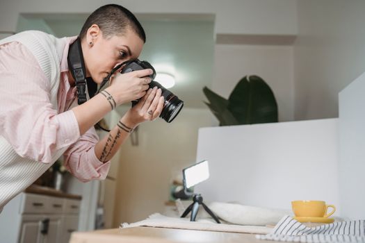 Side view of tattooed woman taking photo of yellow cup on digital camera against table with glowing lamp at home